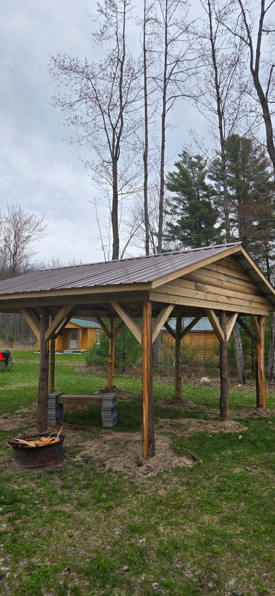 Ein hölzernes Pavillon mit geneigtem Dach, gestützt von Holzbalken, in einer grünen Parklandschaft mit Bäumen im Hintergrund.