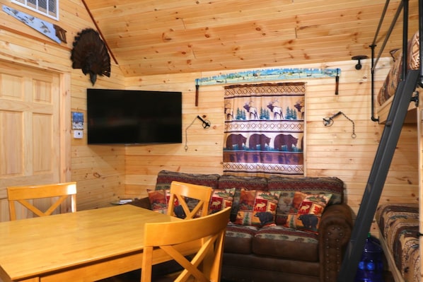 A wooden cabin interior with a dining table, chairs, a sofa with patterned cushions, a mounted television, and a decorative wall hanging. - Cozy cabin located in Brasher Falls, NY. Pet friendly! (Brasher Falls)