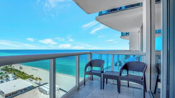 Un balcon avec deux fauteuils en osier, une balustrade en verre et une vue sur une plage et l’océan.
