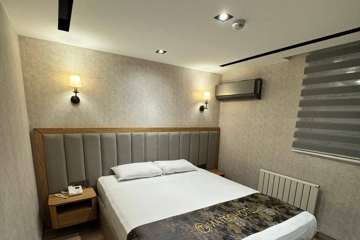 A modern hotel room with a large bed, two bedside tables, a wall-mounted air conditioner, and a window with blinds.