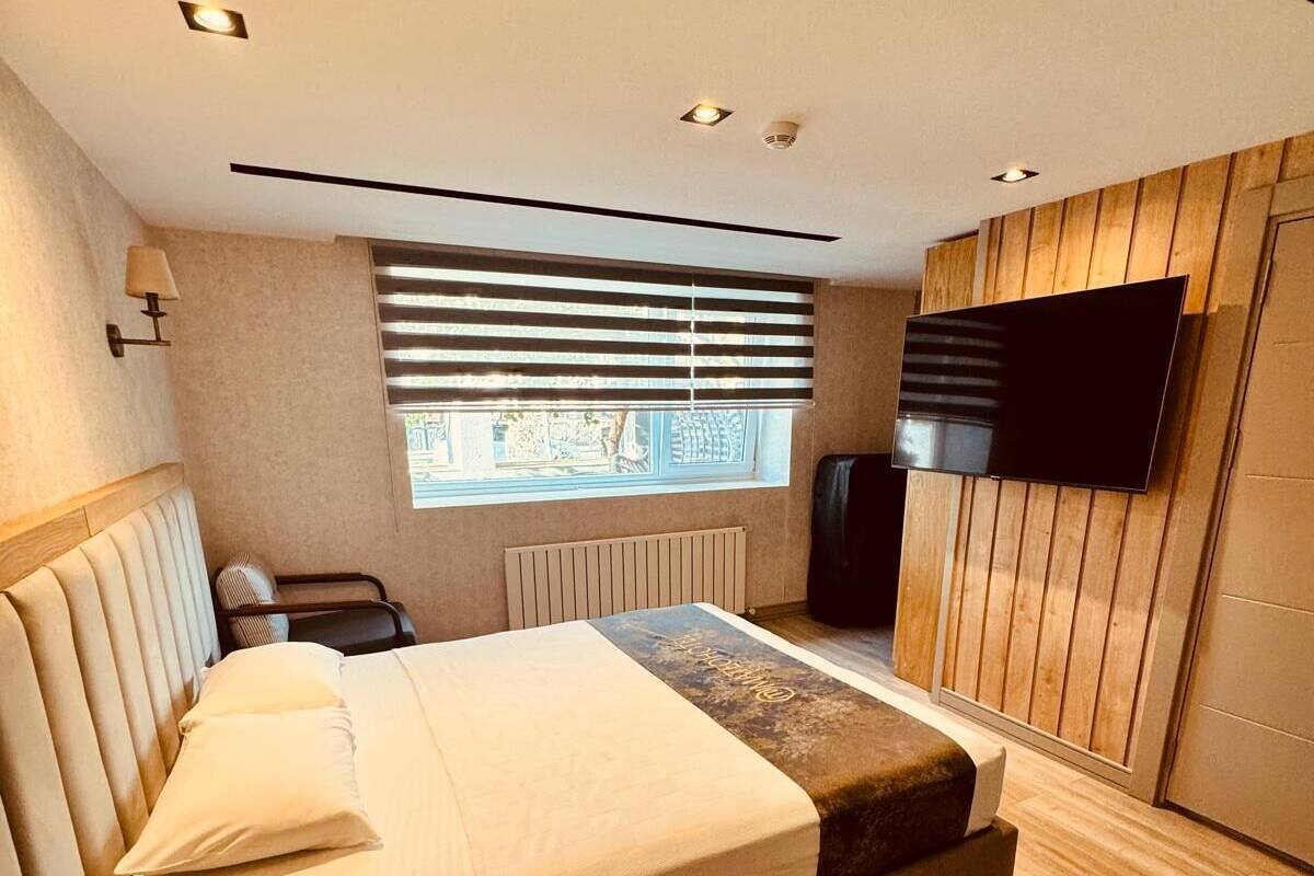 A modern bedroom with a large bed, a bedside telephone, a window with blinds, a radiator, and a mounted television.