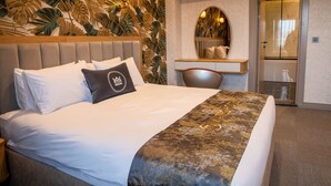 Habitación de hotel con cama, cabecera, pared con estampado de hojas tropicales, espejo y puerta.