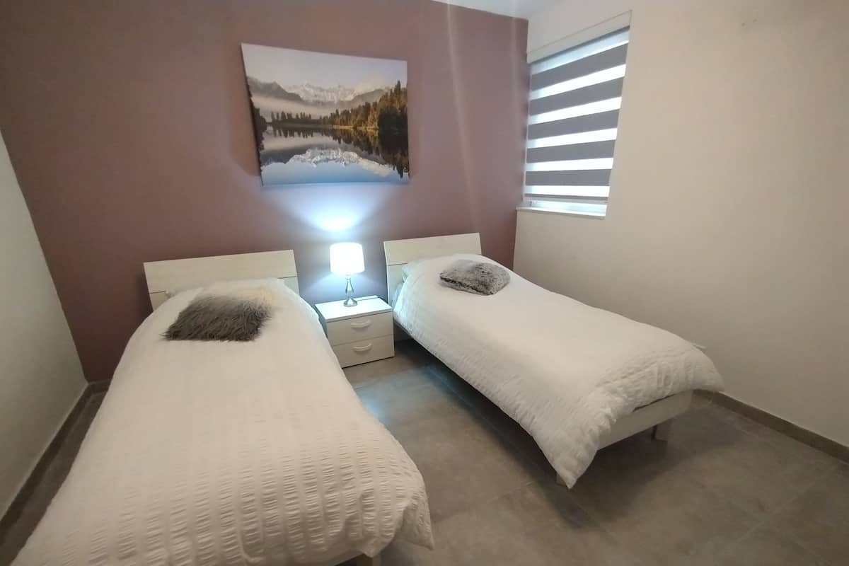 Ein Schlafzimmer mit zwei Einzelbetten, einem Nachttisch mit Lampe, einem Gemälde an der Wand und einem Fenster mit Jalousien.