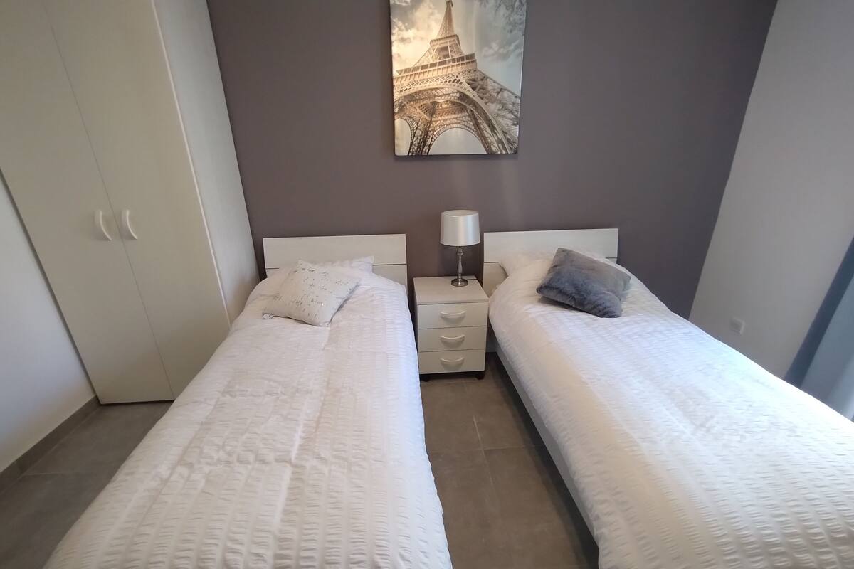 Ein Schlafzimmer mit zwei Einzelbetten, einem Nachttisch mit Lampe, einem Kleiderschrank und einem an der Wand befestigten Bild der Eiffelturms.