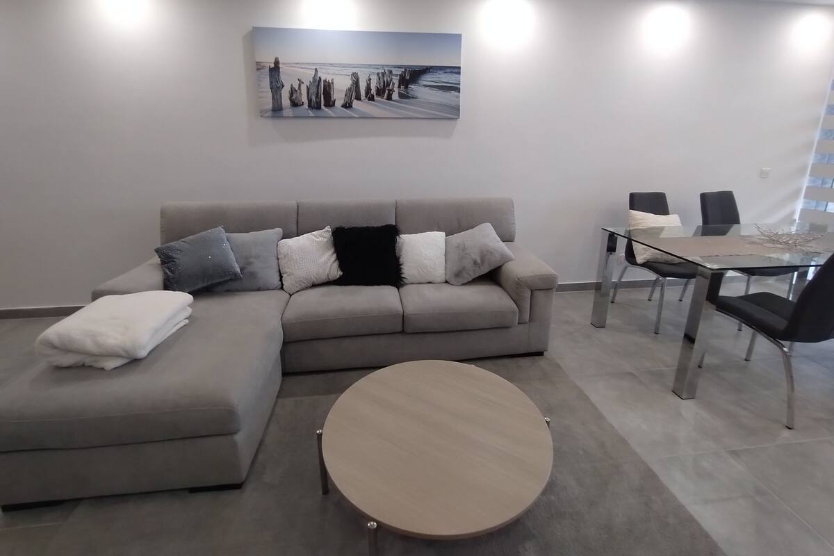Ein modernes Wohnzimmer mit einem grauen Ecksofa, einem runden Couchtisch, einem Glastisch mit Stühlen und einem Gemälde an der Wand.