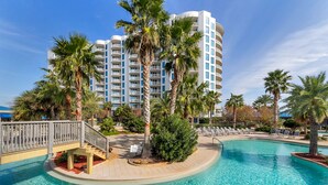 Zona de piscina en un resort, con una piscina circular, palmeras y un edificio de varios pisos al fondo.
