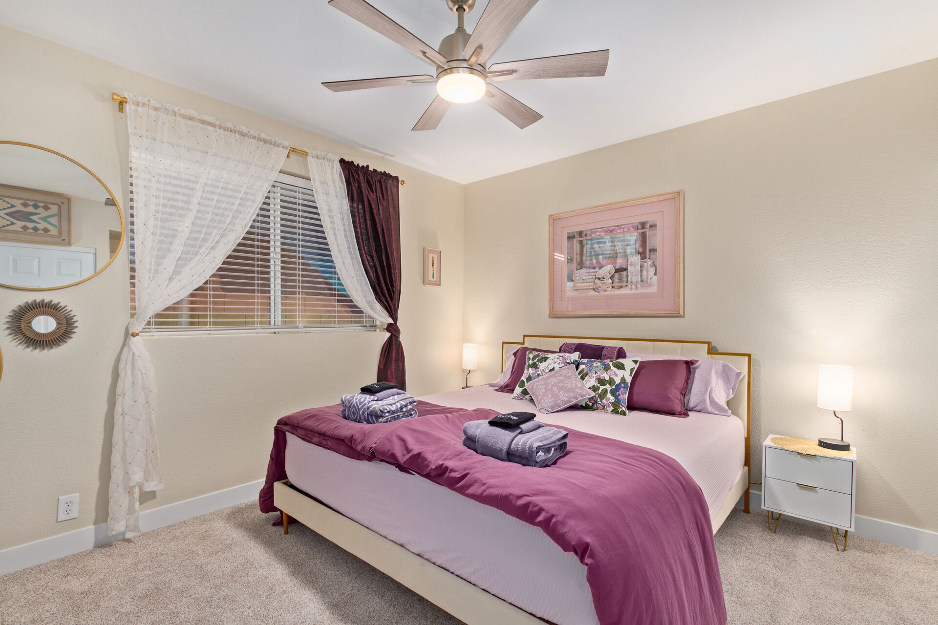 A bedroom with a bed, a nightstand, a ceiling fan, a window with curtains, and wall decorations.