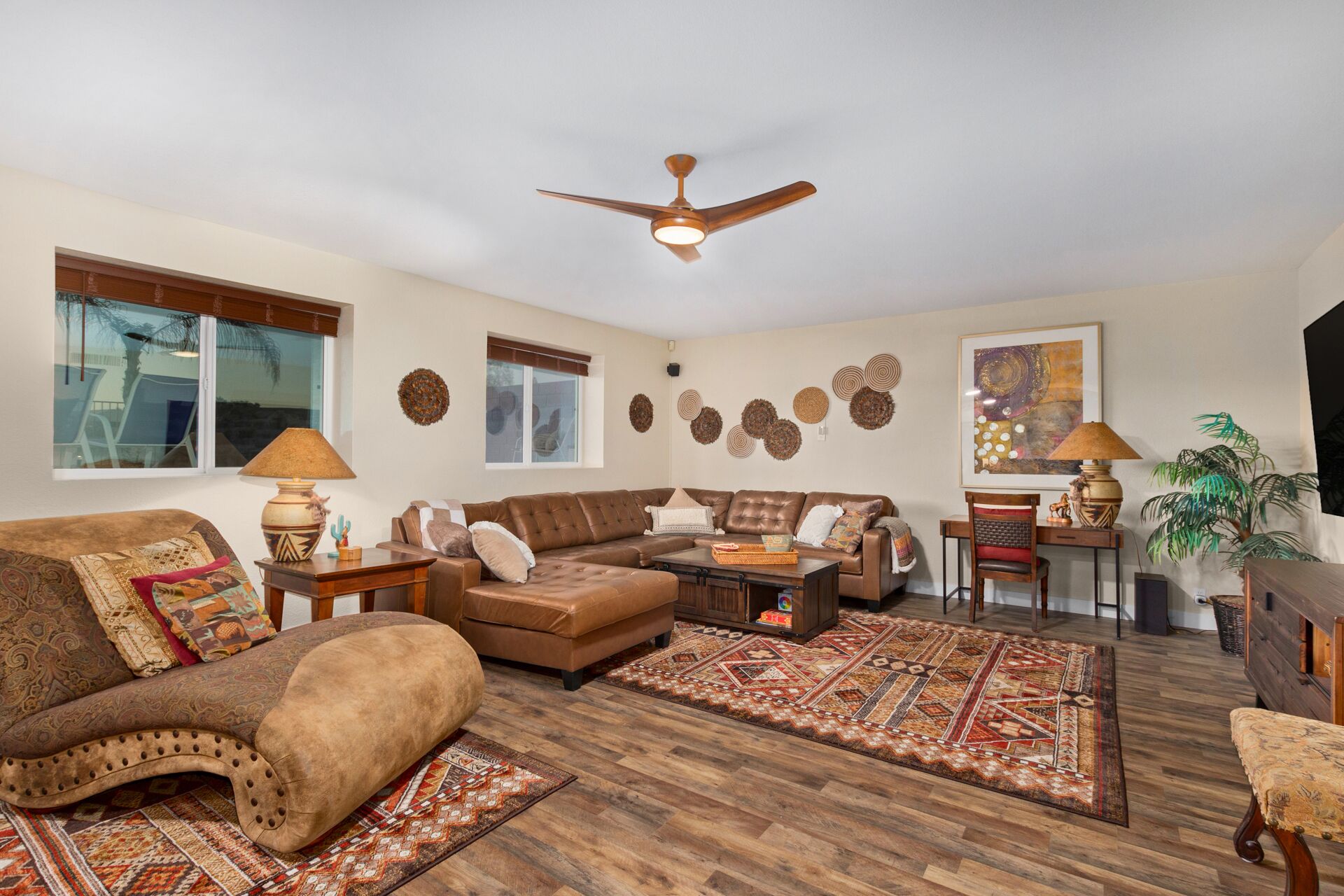 A spacious living room with a brown leather sofa, a wooden coffee table, and a patterned rug. There is a ceiling fan, a side table with a lamp, and a wall-mounted lamp.