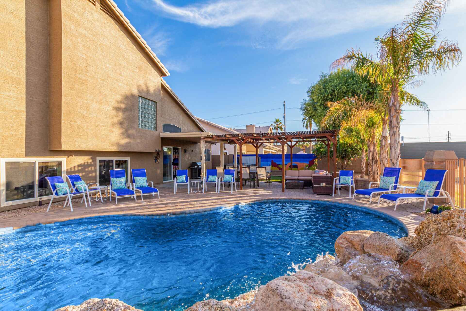 A swimming pool with lounge chairs and a pergola, surrounded by a building with large windows and a view of palm trees.