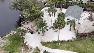 A house with a deck overlooking a body of water, surrounded by palm trees and a sandy area.