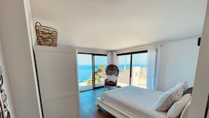 Ein Schlafzimmer mit Bett, Sessel, Nachttisch und Meerblick durch große Fenster.