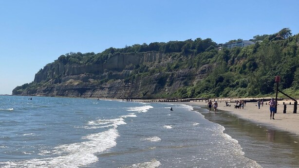 Une plage avec une falaise rocheuse en arrière-plan, une promenade en bois longeant le rivage, et des gens qui se promènent et qui nagent.