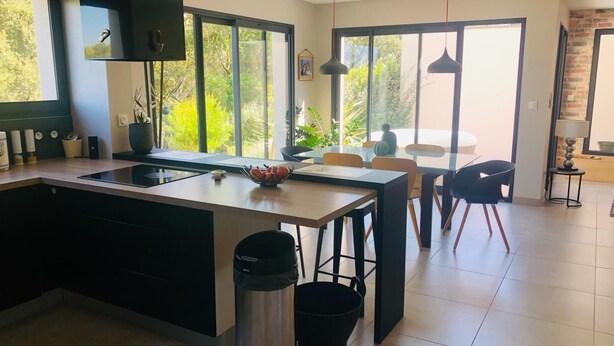 Une cuisine moderne dotée d’un îlot central, de grandes fenêtres et d’un coin repas avec des chaises et une table.