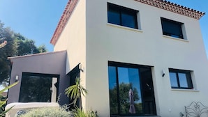 Une maison moderne à deux étages, dotée de grandes fenêtres, d’un toit en tuiles et d’un jardin agrémenté de plantes et d’un banc.
