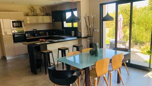Une cuisine moderne avec un îlot central, des tabourets de bar noirs, une table à manger en verre avec des chaises jaunes, et une vue sur la verdure grâce à de grandes fenêtres.