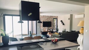 Une cuisine moderne dotée d’une cuisinière intégrée, un coin repas avec une table en verre et un salon avec un canapé et un téléviseur fixé à un mur en briques.