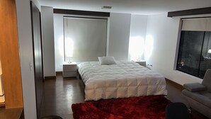 A bedroom with a bed, a nightstand, a red rug, a window with a view, and a chair. - Departamento Amoblado Lloyd Apartment 94 (Bogotá)