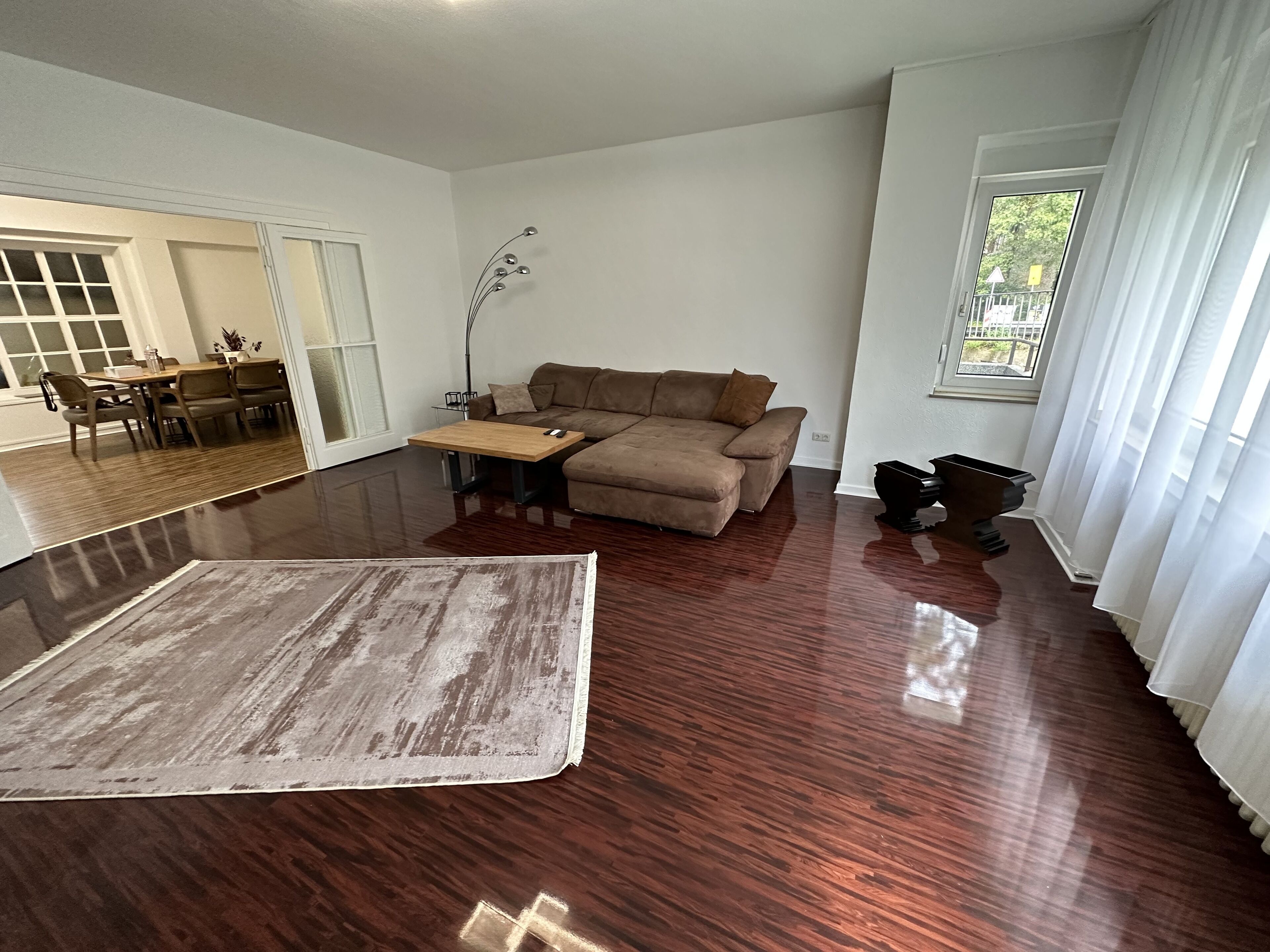 Un salon spacieux doté d’un grand plancher en bois, d’un canapé marron, d’une table à manger avec des chaises et d’une grande fenêtre agrémentée de rideaux.