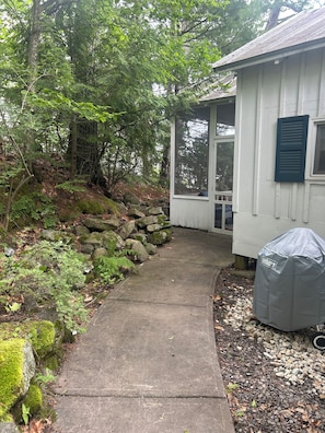 A concrete pathway leading to a white building with a covered porch, surrounded by trees and rocks.