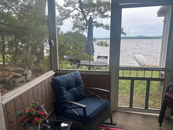 A covered porch with a blue cushioned chair, a small table with chairs, and a view of a lake.