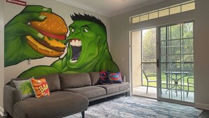 A living room with a large mural of a green muscular figure holding a burger, a gray sofa, and a glass door leading to a patio.