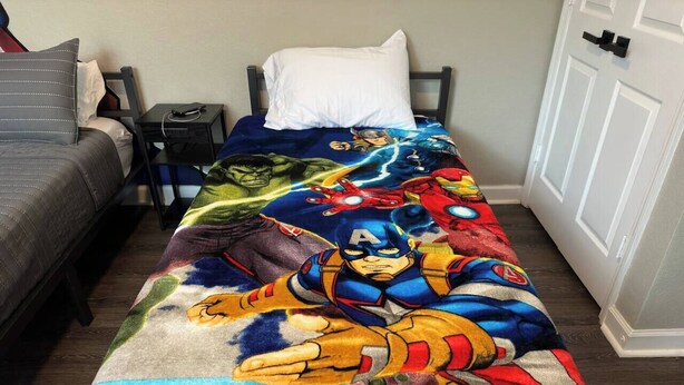 A bed with a superhero-themed blanket, a bedside table with a phone, and a nightstand with a lamp.