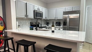 A kitchen with white cabinets, a stainless steel refrigerator, a microwave, and a coffee maker. There are three black stools at the counter.