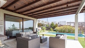 Un espacio de estar al aire libre techado con mobiliario de mimbre, una puerta de cristal que da a una piscina y vistas a una zona residencial al fondo.