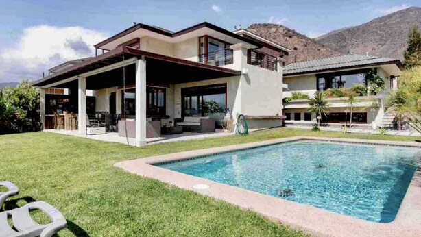 Una casa de dos pisos con piscina, un porche cubierto y montañas al fondo.