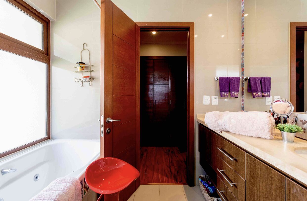 Une salle de bain moderne avec une baignoire, une porte en bois donnant sur une pièce sombre, un meuble-lavabo avec un miroir et une chaise rouge.