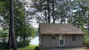 A small, single-story house with a sloped roof, a front door, and two windows. It is surrounded by a grassy yard and trees. - Brimfield Waterfront cottage (Brimfield)