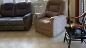 A living room with a gray sofa, a beige armchair, and a wooden rocking chair. There are large windows allowing natural light. - Brimfield Waterfront cottage (Brimfield)