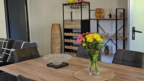 Ein Essbereich mit einem Holztisch, grau gepolsterten Stühlen, einer Vase mit gelben Blumen und einer schwarzen Regaleinheit mit Dekorationen.