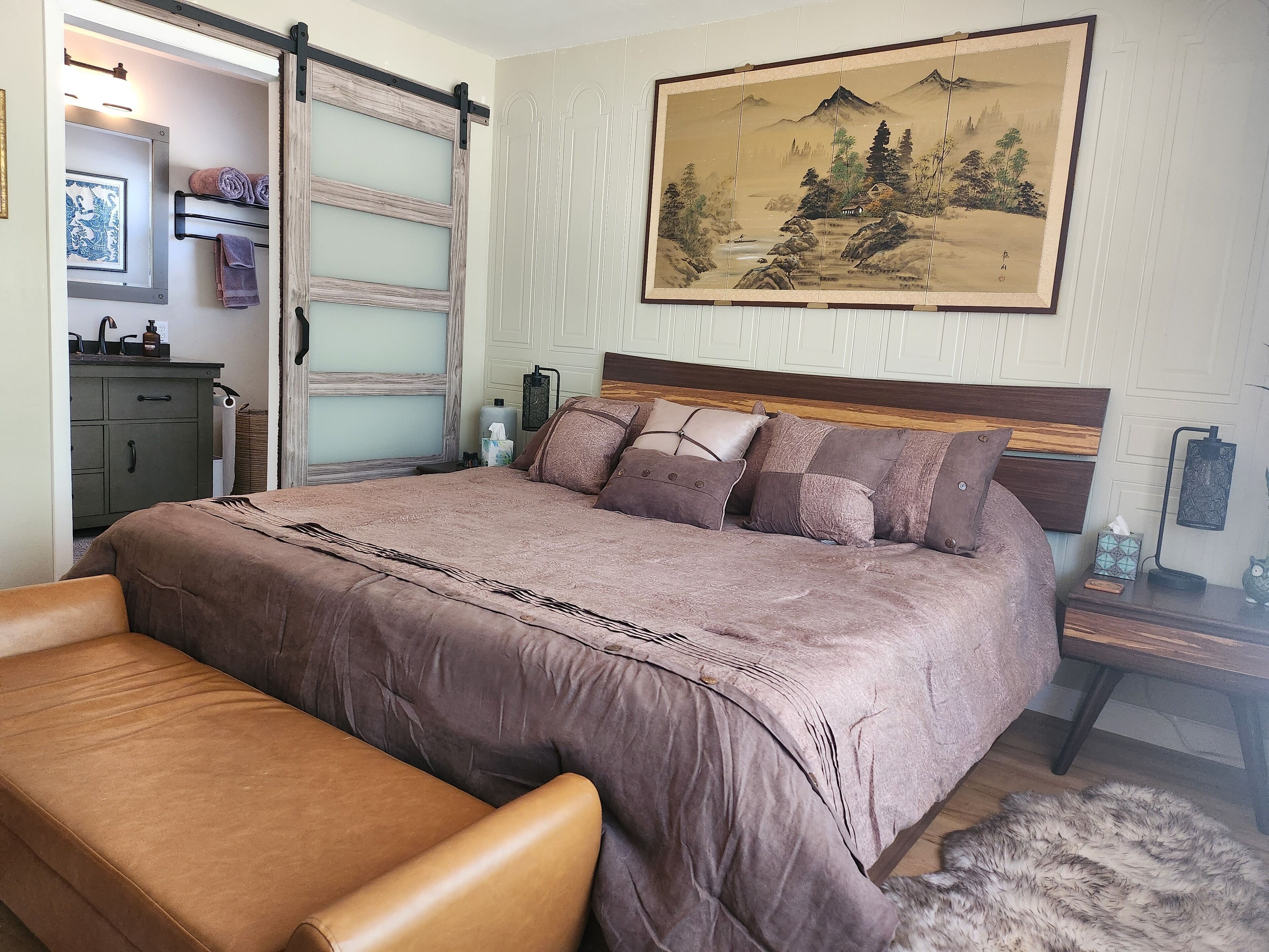 A bedroom with a large bed, a bench, a dresser, a mirror, and a painting on the wall.