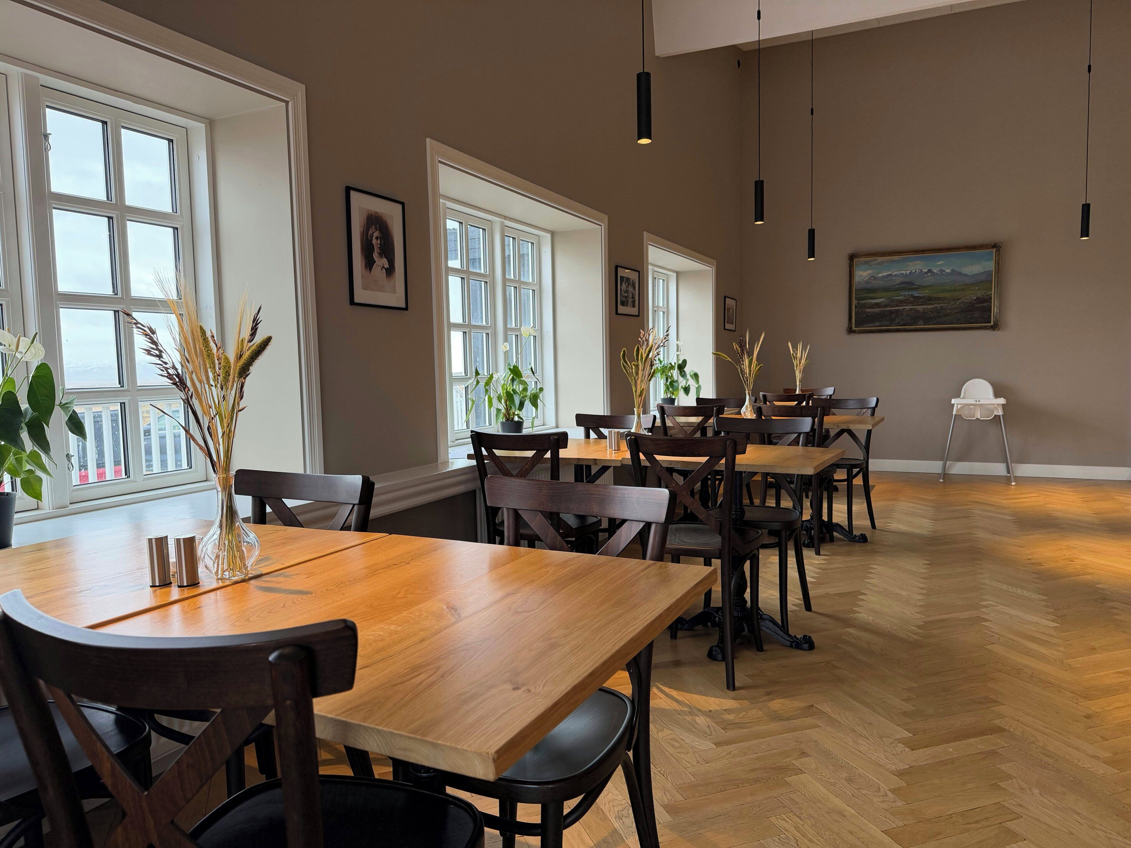 A dining area with wooden tables and chairs, large windows, and hanging pendant lights.