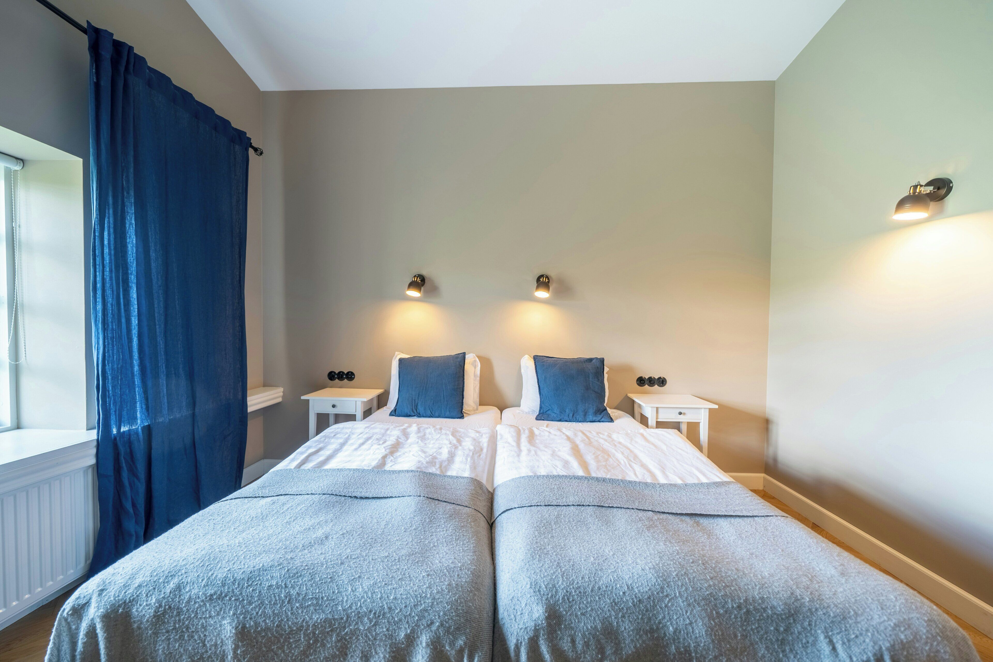 Habitación doble con dos camas, ventana con cortinas, mesitas de noche e iluminación en la pared.