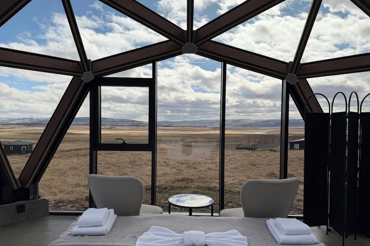 A room with a bed, two chairs, a small table, and a large window offering a view of a landscape.