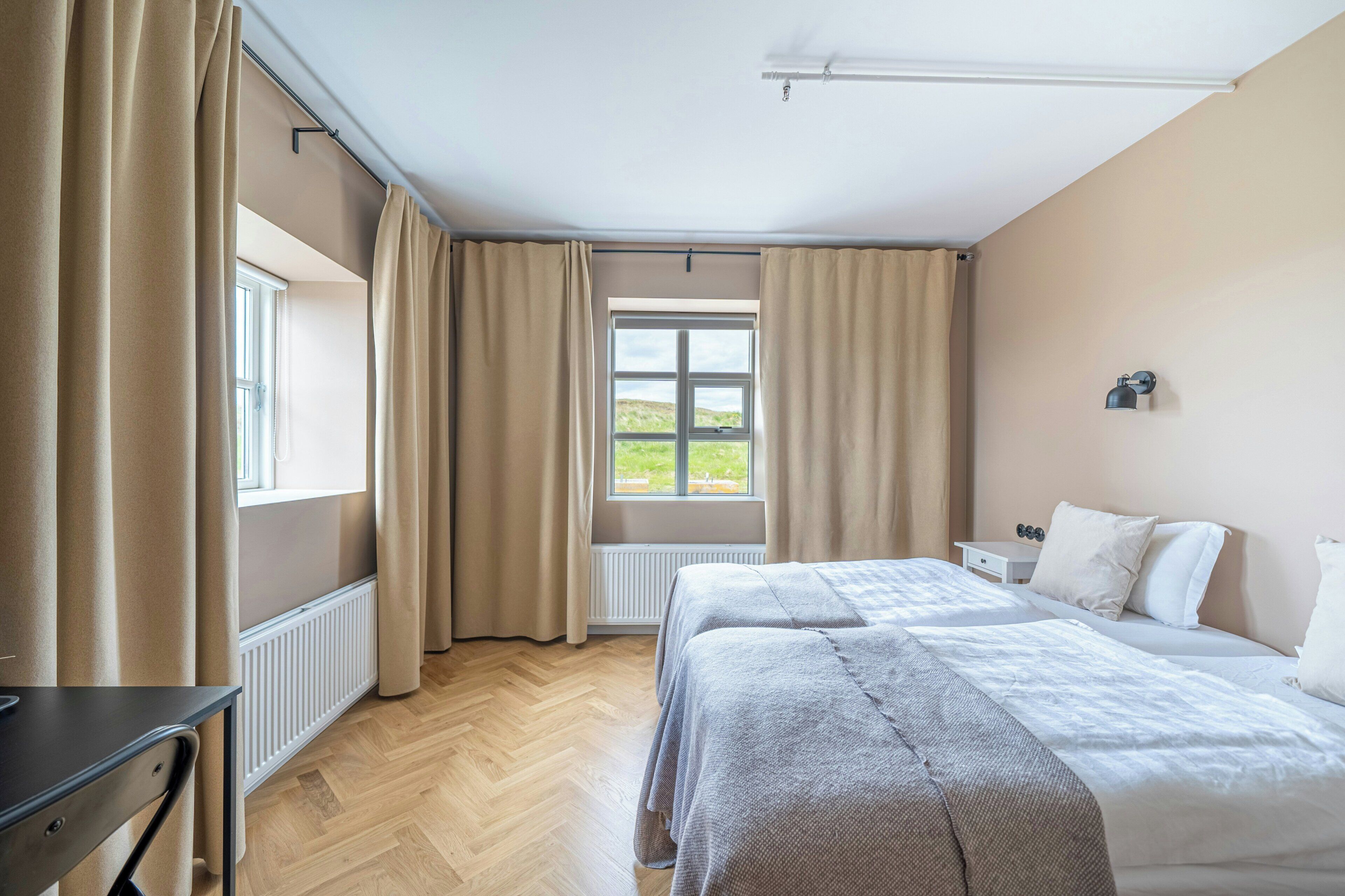 A hotel room with two beds, a window with curtains, a radiator, and a small table with decorative items.