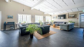 A spacious living room with dark flooring, a sectional sofa, a grey armchair, and a coffee table. There are large windows allowing natural light, a ceiling with a grid pattern, and a potted plant in the center. - Hotel Stafholt (Borgarbyggd)