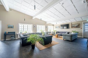 Amplia sala de estar con piso oscuro, sofá modular, un sillón gris y una mesa de centro. Hay ventanas grandes que dejan entrar luz natural, un techo con diseño de cuadrícula y una planta en maceta en el centro.