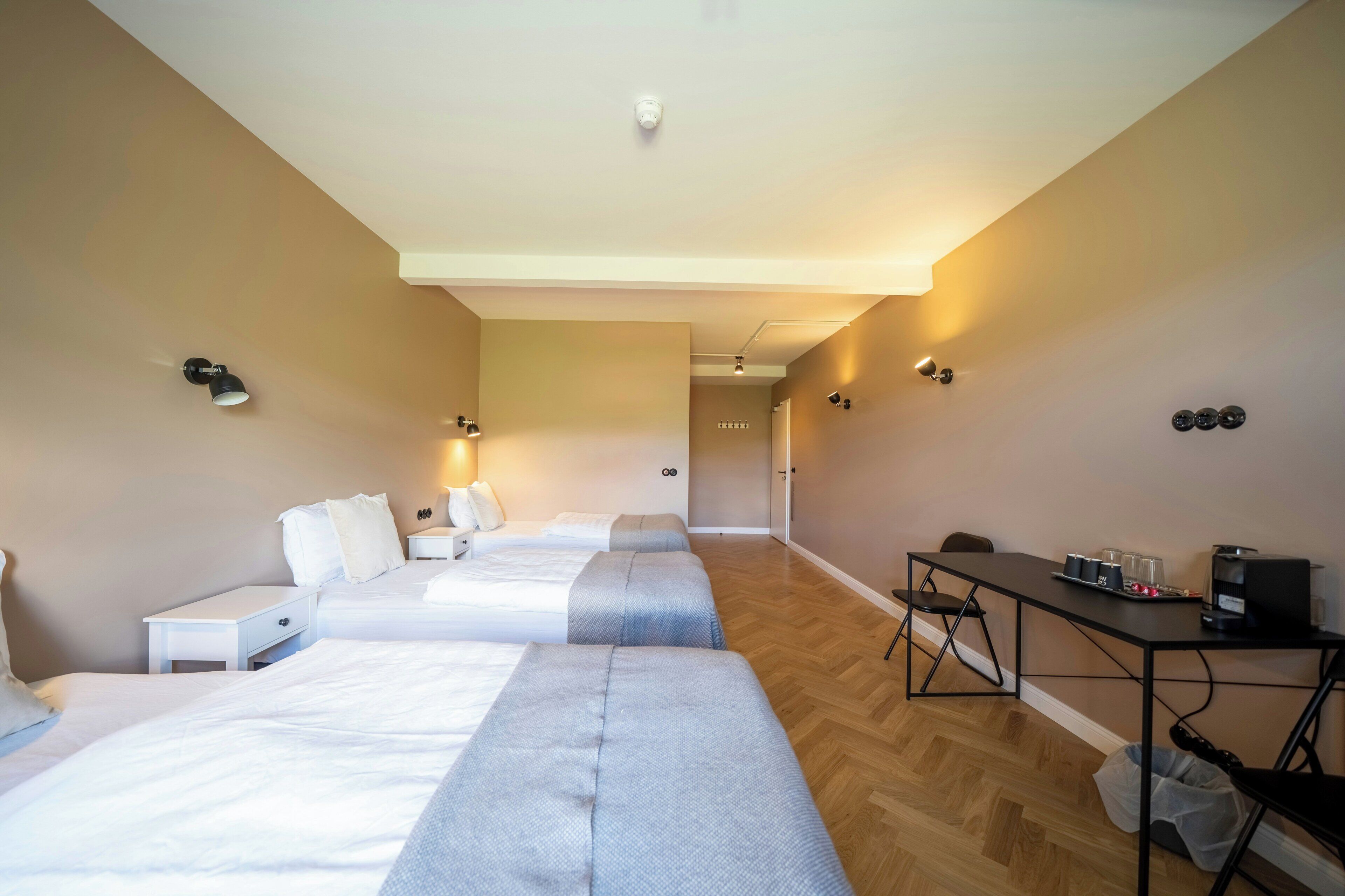 Une chambre d’hôtel avec trois lits, un bureau avec une machine à café et une petite table avec des tasses et des verres.