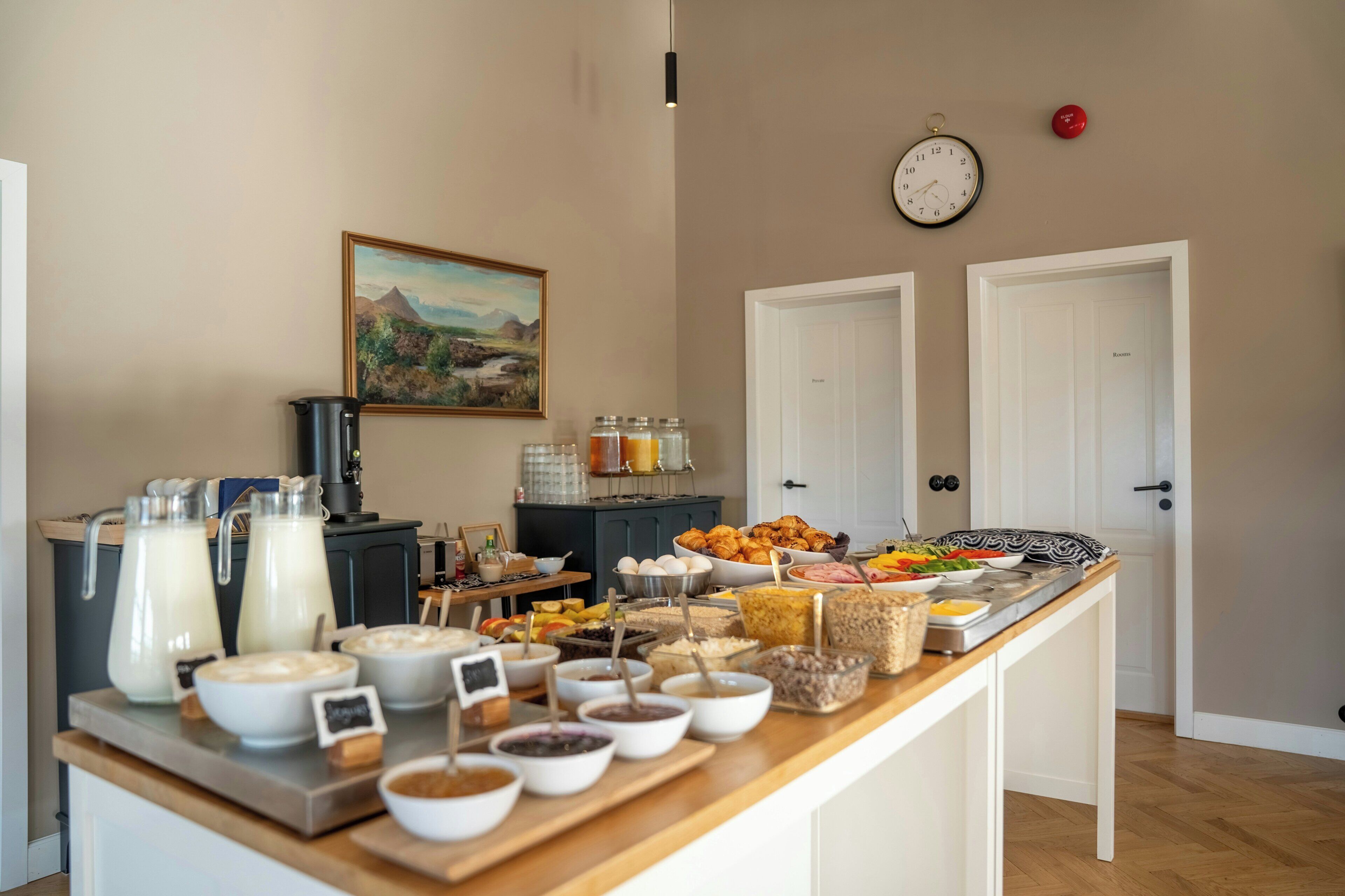 A breakfast buffet with various food items and beverages, a clock, and a painting on the wall.