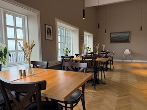 A dining area with wooden tables and chairs, large windows, and hanging pendant lights. - Hotel Stafholt (Borgarbyggd)