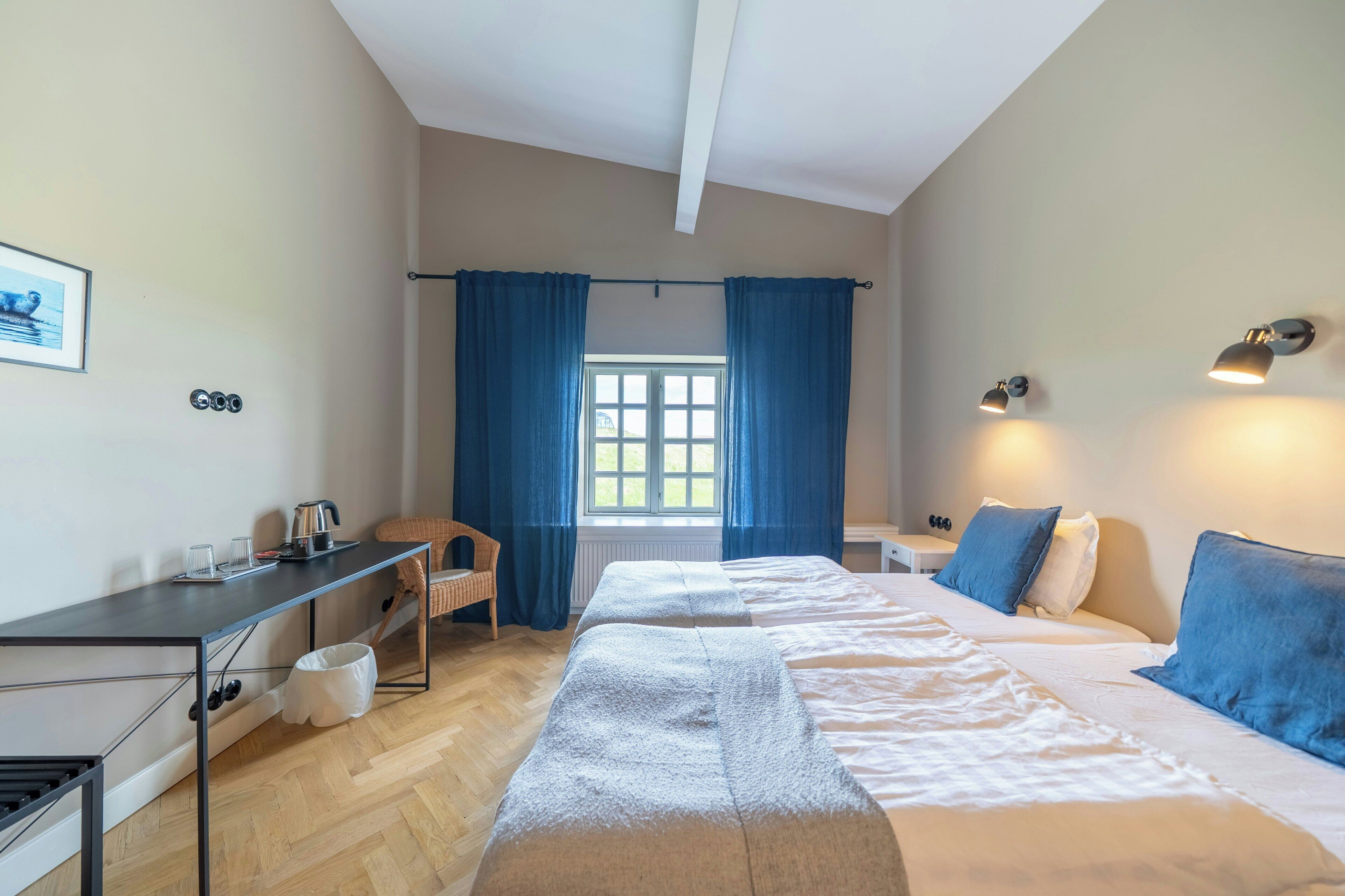 Habitación de hotel con cama, escritorio, silla, ventana con cortinas y lámparas fijadas a la pared.