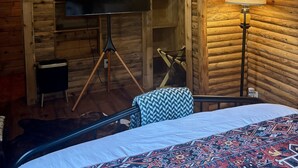 Una habitación estilo cabaña de madera con una cama, un televisor sobre un trípode, un ventilador de techo y una lámpara.