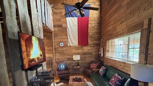 Un interior de cabaña acogedor con paredes de madera, un ventilador de techo, una bandera, una chimenea, un sofá y una mesa.