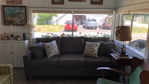 A living room with a dark grey sofa, patterned cushions, a wooden side table with a lamp, and a view of parked cars outside. - Cozy beach cottage. Less than a 2minute walk to the beach.  (Westbrook)