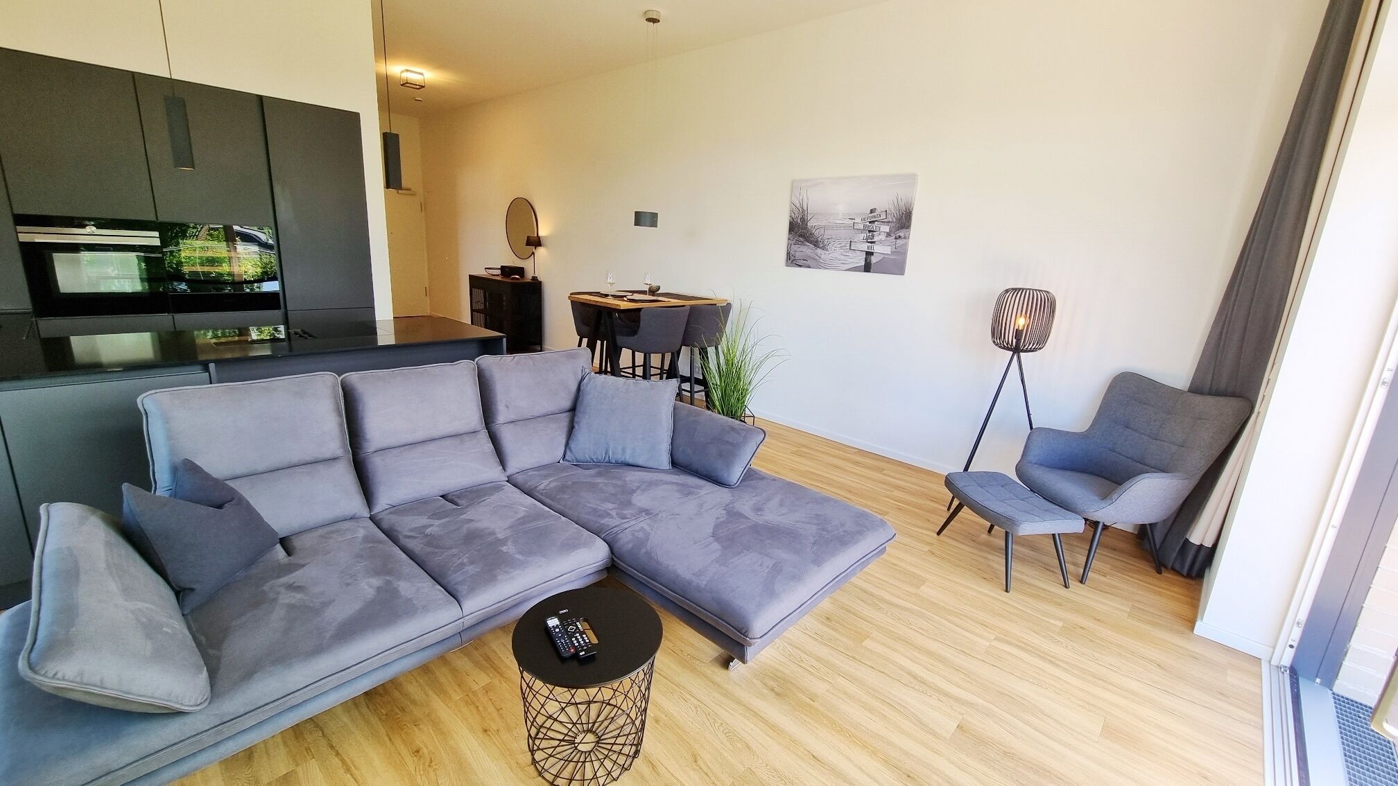 Ein modernes Wohnzimmer mit einem grauen Ecksofa, einem kleinen runden Couchtisch und einem grauen Sessel. Es gibt eine Essecke mit Tisch und Stühlen sowie eine Stehlampe.