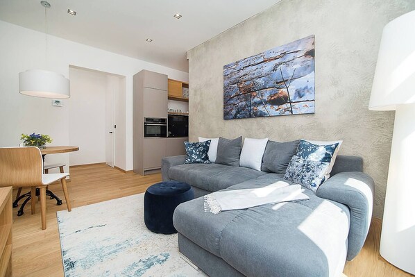 A modern living room with a grey sofa, a round ottoman, a dining table with chairs, and a kitchen in the background.