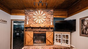 A rustic fireplace with a brick surround and wooden mantels, a television mounted above it, and a kitchen visible in the background.